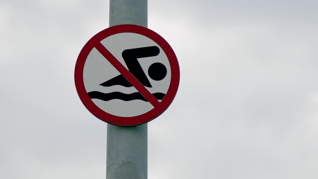 Close-up static shot of a no swimming prohibition sign mounted on a metal pole against an overcast sky. Safety warning symbol indicating swimming is forbidden near water. Minimal outdoor composition.
