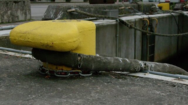 Close-up of a yellow mooring bollard with heavy marine rope secured on a harbor dock. Static shot highlighting maritime infrastructure, port equipment and nautical details beside a working marina.