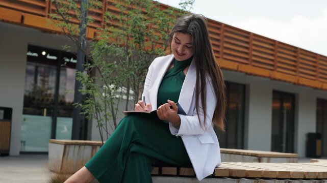 Beautiful young business woman sitting on wooden bench and writing in notebook