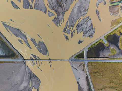 Aerial view of a bridge crossing a complex braided river system with sediment-rich glacial water and surrounding volcanic terrain in Iceland.
