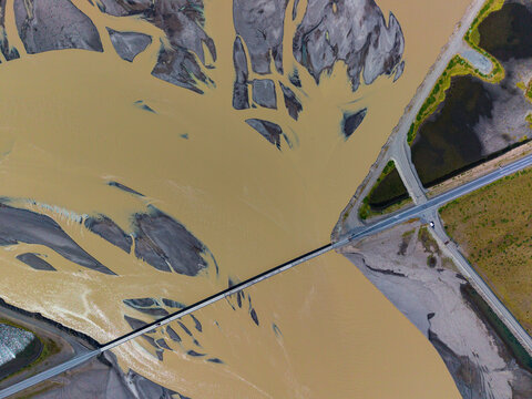 Aerial view of a bridge crossing a wide braided river delta with sediment-rich glacial water and sparse vegetation in Iceland.