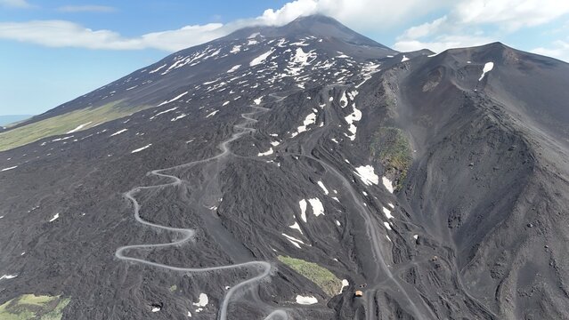 Aerial view of Mount Etna featuring a winding road traversing the volcanic landscape with patches of snow and dark lava rock in Castiglione di Sicilia, Sicily, Italy.