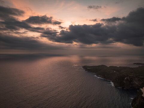 Aerial view of the rugged coastline of Penida Island at sunset with dramatic clouds and golden light reflecting on the ocean in Nusa Penida, Bali, Indonesia.