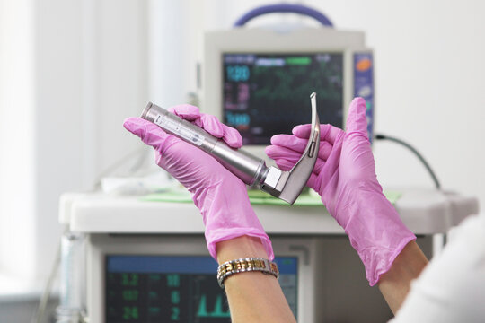 Doctor's hands in pink nitrile gloves hold medical laryngoscope against background of heart monitor in operating room