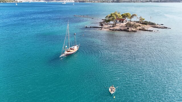 Aerial view of the small island of Poros with a white chapel and pine trees, a sailboat anchored in the turquoise sea, and a person rowing a small wooden boat in Poros, Greece.