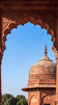 Intricately carved red sandstone archway framing an ancient domed monument against a clear blue sky in India