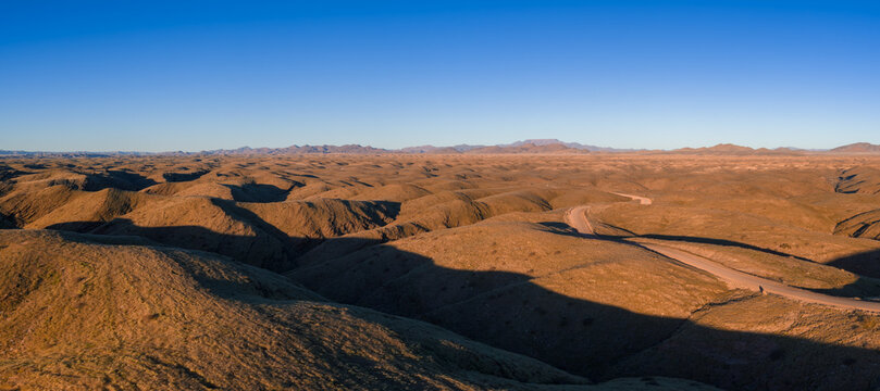 Aerial view of rolling ochre hills, dry ravines, and a lone dirt road in Namibia near the Namib Desert, late afternoon light casts long shadows under a clear sky.