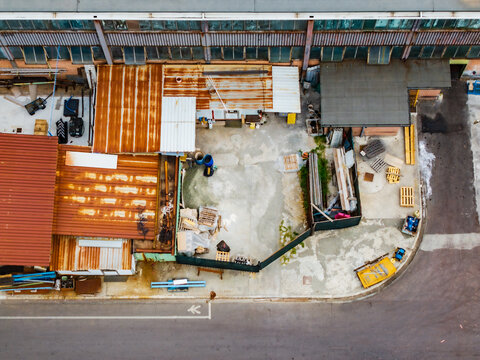 Aerial view of the Serravalle Logistic area featuring industrial buildings with rusted roofs, storage yards with wooden pallets, and an asphalt road in Serravalle Scrivia, Piedmont, Italy.