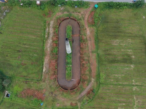 Overhead top-down drone shot showing a uniquely shaped oval dirt track or ground pattern surrounded by green paddy fields and countryside.