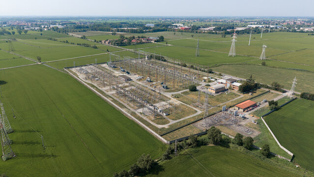 Aerial view of the C.P. Lacchiarella electrical substation surrounded by green agricultural fields, power lines, and metal pylons in the rural landscape of Badile, Lombardia, Italy.