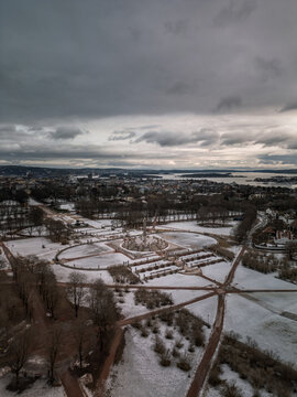 Aerial view of Frogner Park in winter with snow-covered grounds, the Monolith sculpture, and the surrounding city of Oslo under a cloudy sky, Oslo, Oslo, Norway.