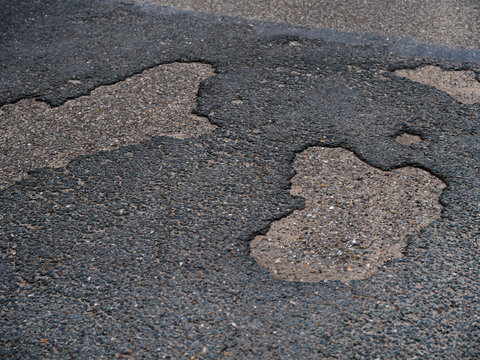Large potholes and cracks in a worn asphalt road surface reveal the gravel beneath.