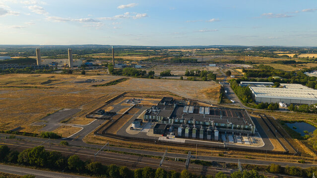 Aerial view of a modern hyperscale data center facility surrounded by industrial land, railway tracks, and open fields during golden hour in Didcot, England, United Kingdom.