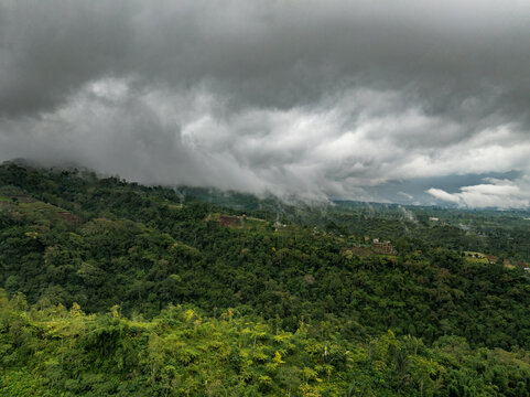 Aerial view of the lush green hills and forests of Bedugul under a dramatic, heavy overcast sky with low-hanging clouds in Kecamatan Baturiti, Bali, Indonesia.