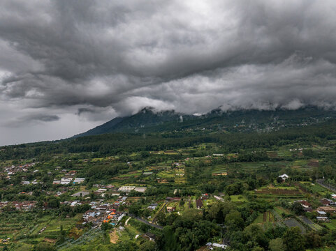 Aerial view of Bedugul with lush terraced fields, a small town, and dramatic storm clouds over the mountains, Kecamatan Baturiti, Bali, Indonesia.