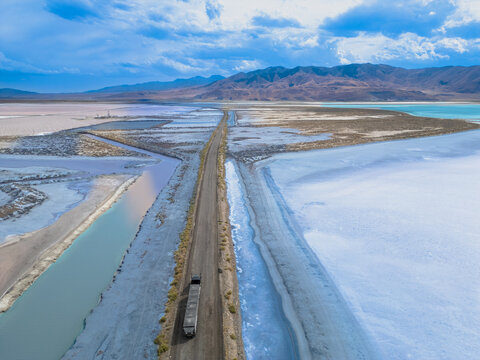 Aerial view of a truck driving along a narrow road through the colorful salt flats and evaporation ponds with distant mountains under a cloudy sky in Grantsville, Utah, United States.