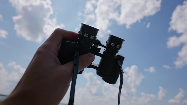 A hand holds black binoculars up towards a bright blue sky filled with fluffy white clouds. Concept of exploration, outdoor adventure, searching for answers, and future vision.