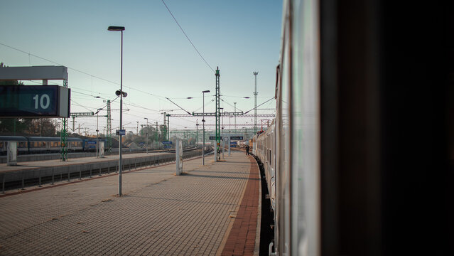 A train platform at Kelenfold stretches beneath overhead catenary wires and signal poles at a busy railway station, with a passenger train waiting alongside under a clear morning sky.