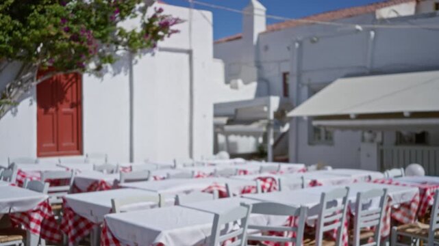 Blurred outdoor cafe terrace in mykonos with soft whitewashed walls, red checked tablecloths and stacked chairs; background backplate copyspace calm.
