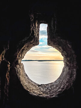 Scenic sea view through a medieval stone loophole with warm sunset light reflecting on calm water. Historic defensive architecture overlooking the coastline.