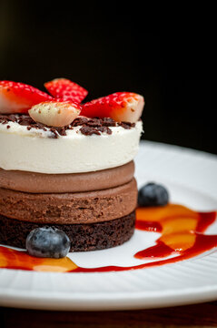 Close up of layered mousse cake with strawberries. Fresh sliced berries sit on top of the dessert drizzled with fruit sauce on a ceramic plate.