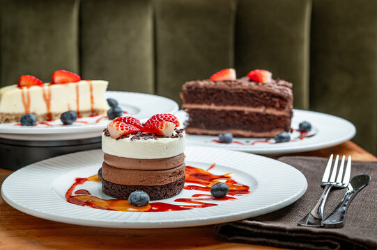 Tasty desserts on cafe plates. Three different cakes are displayed on white plates at a cafe. The cakes have fruits and sauces on top.