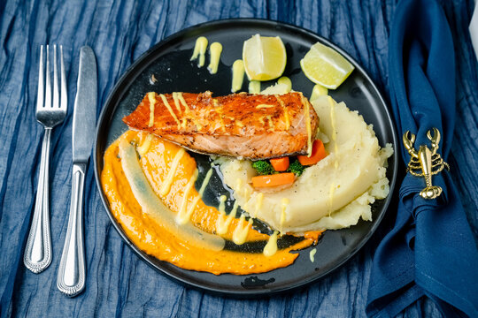 Salmon dish served with mashed potatoes. A plate shows salmon fillet on mashed potatoes with vegetables and sauce. Lime wedges are placed beside the food.