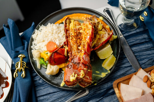 Cooked lobster tail platter with rice and vegetables. A whole lobster tail is presented on a dark plate with rice, mixed vegetables, and lemon slices.