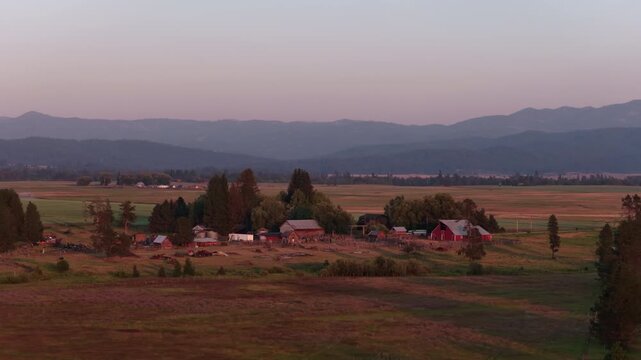 Aerial of rural farmstead and valley floor at sunset in Donnelly Idaho with multiple barns and pink glowing sky with wildfire smoke in summer