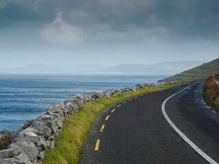 Asphalt road curves beside endless ocean. Grass grows between stones and pavement. Sky holds soft clouds above the sea. Road leads toward distant horizon. Perfect for travel, nature, or coastal themes © mark_gusev