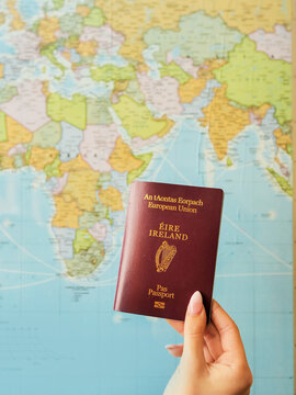 Woman holding passport of Ireland in her well manicured hand, world map in the background with. Emigration of young Irish people abroad from Ireland due to high cost of living concept.