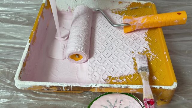 A person's hand rolls a paint roller with an orange handle back and forth on the textured ramp to evenly distribute the paint. A home painting