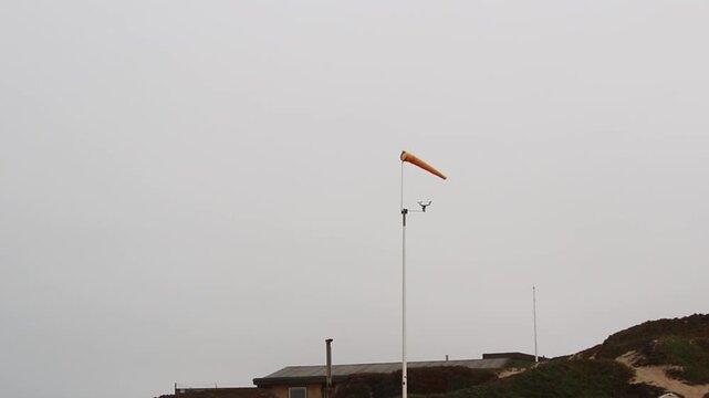 Orange Windsock And Cup Anemometer Spinning On Top Of Pole Against Overcast Sky With Sand Dune And Building Roof
