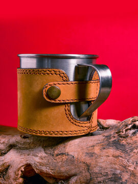 Stainless steel mug wrapped in brown leather sleeve. Mug rests on weathered wooden log. Vibrant red background behind. Leather strap with metal button closure