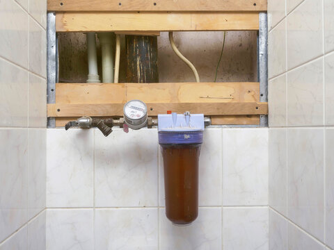 Water filter unit and meter mounted on tiled wall. Pipes visible behind wooden frame. Brownish water inside filter housing. Metal fittings and pressure gauge attached. Home plumbing installation