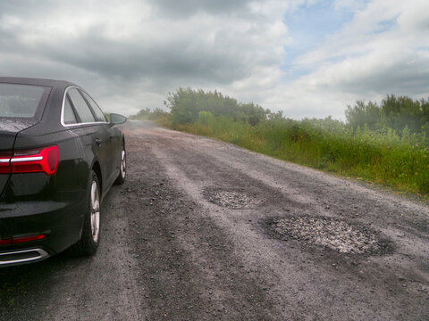 Black car on rough, potholed road. Misty weather, overcast sky. Green vegetation lines the roadside. Road stretches into distance, blurred by fog. Living in remote country side with bad road surface