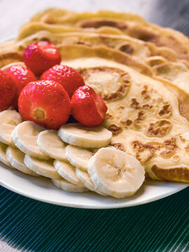 Stack of golden-brown pancakes on white plate. Fresh strawberries and sliced bananas beside pancakes. Soft and dreamy look, Hand made simple country meal. Dark wooden surface background