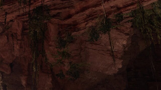 In a dark cave, there are large rock formations with moss and roots hanging down. Light filters through cracks above, illuminating the cave walls. The space is cool and damp.