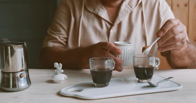 Man making coffee at home, putting cane sugar into glass coffee cup with hot coffee. Preparation home coffee, morning ritual, coffee brewing process.