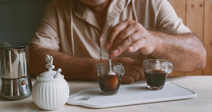 Man making coffee at home, putting cane sugar into glass coffee cup with hot coffee. Preparation home coffee, morning ritual, coffee brewing process.
