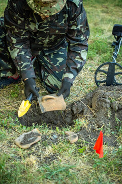 A archaeologist in camouflage clothes uses a metal detector in a grassy area. They are digging and finding some items in the soil. Red flags mark the spots where they search