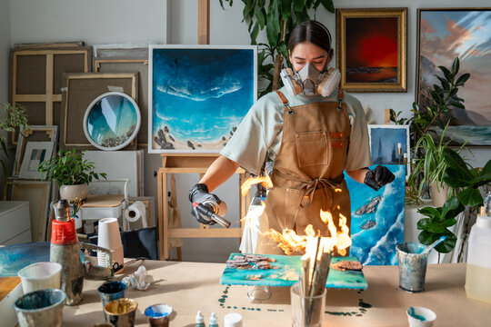 Woman crafter in respirator using professional tools and creating liquid art using epoxy resin, craftswoman artist using gas torch to heat top layer of uncured epoxy to remove air bubbles from surface
