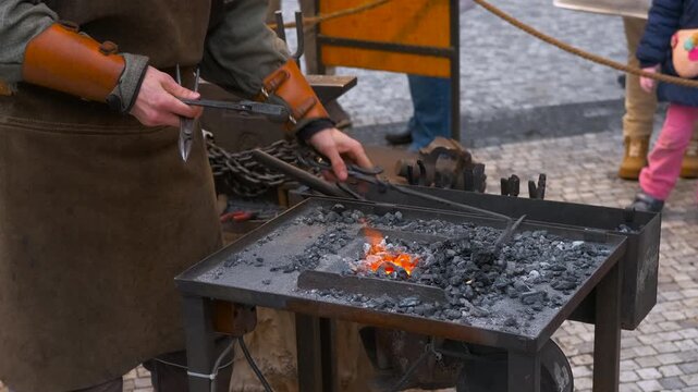 Blacksmith forging metal in hot coals with sparks flying. Professional blacksmith in a leather apron heating a piece of iron in a traditional coal forge, using tongs to manipulate the glowing metal