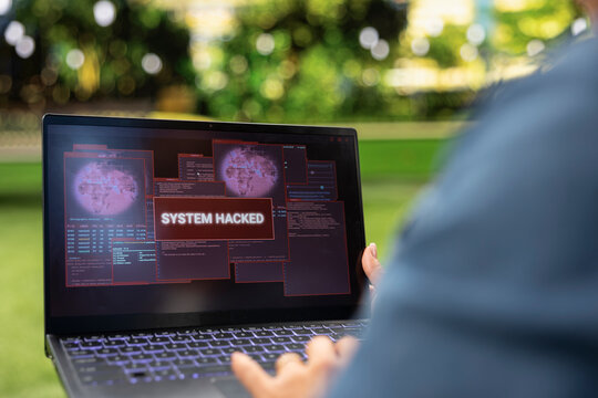 Close up of a laptop showing a red system hacked warning and digital threat while a person works outdoors, illustrating cyberattack crime, malware risk and a compromised computer in a city park.