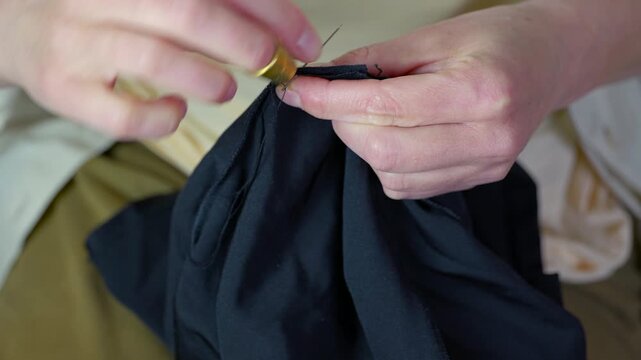 Clothing repair. Female tailor's hands meticulously sewing black fabric with a needle and thread, using a thimble for protection. Skillful dressmaker is repairing or creating handmade clothes