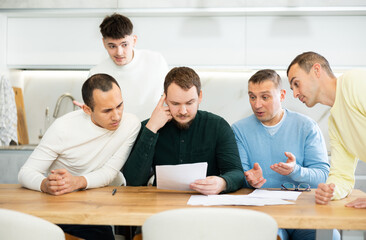 Fototapeta premium Upset worried man sitting at table in home kitchen with papers in hands, reading unexpected medical results with group of male friends solacing and comforting him ....