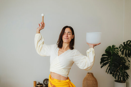 Relaxed woman balances a singing bowl and mallet in a bright home, natural photographic style, wellness meditation concept
