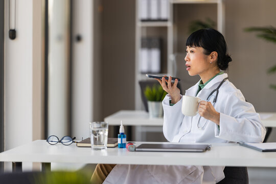 Female doctor recording voice message on smartphone while holding coffee in clinic office