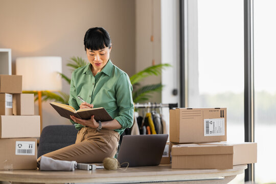 Female entrepreneur packing orders at home office for online shop with laptop and cardboard boxes