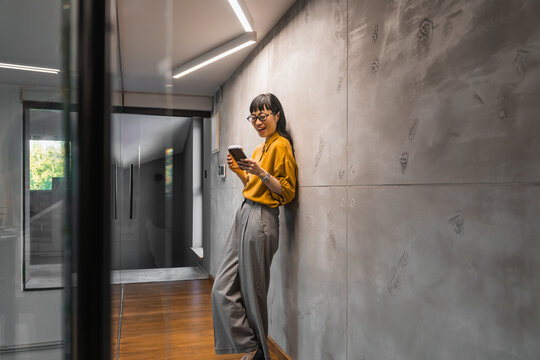 Young businesswoman standing in modern office hallway smiling while using smartphone in casual work attire